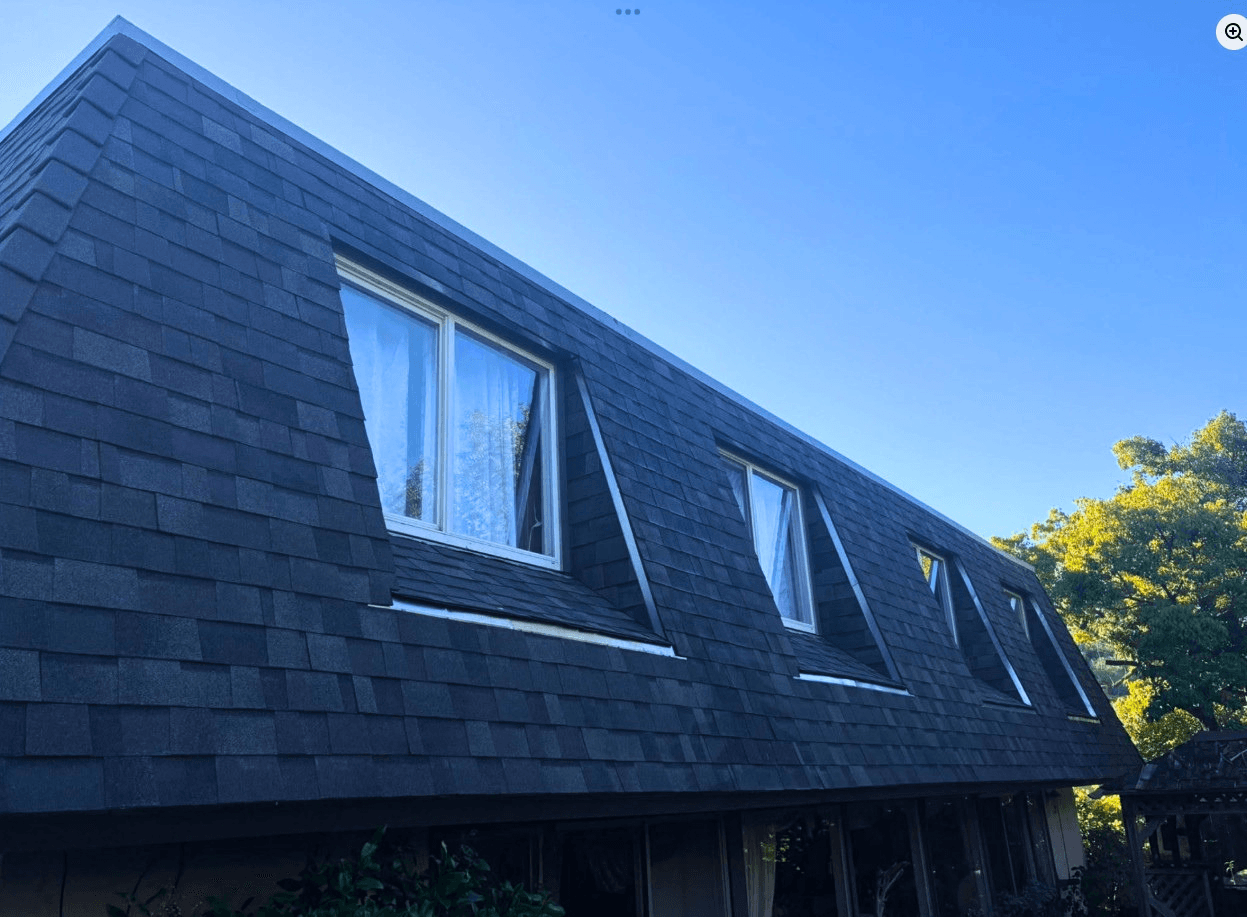 Dark grey shingled mansard roof with four recessed windows set against a clear blue sky.